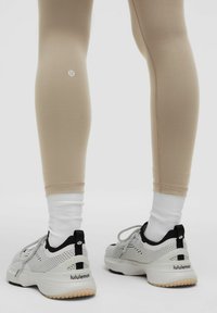 Leggings beige realizzati in tessuto elasticizzato con un design aderente, abbinati a sneakers bianche e nere con dettagli in rete e suola in gomma.
