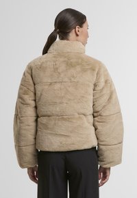 Bež, kunstlikust karusnahast puffjakker kõrge kaelusega, laiade käiste ja tikitud tekstuuriga. Lühike pikkus ja laheda lõikega vöökoht.