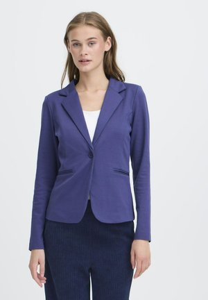 Jonge vrouw in een getailleerde paarse blazer over een witte top en donkere marineblauwe broek, staande tegen een effen witte achtergrond.