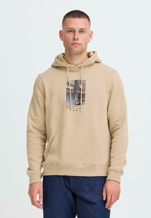 Beige hoodie made of soft fabric with a kangaroo pocket, drawstrings, and a front graphic print. Worn with dark blue jeans.