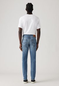 Jeanși din denim albastru deschis, cu un design cu picioare drepte, buzunare spate și un petic din piele maro la talie, asortați cu un tricou alb.