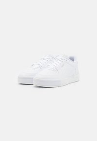 Ett par vita lågtoppade Puma-sneakers med perforerad tåbox, snörning och texturerad sula på en vit bakgrund.