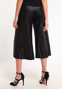 Culottes en simili cuir noir avec un design à jambes larges, texture lisse et détails de couture au centre au-dessus des chevilles. Associé à des talons aiguilles noirs avec des lanières.