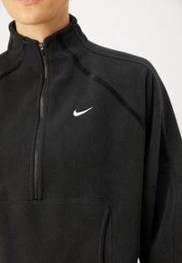 Giacca in pile nera con zip a quarto, caratterizzata da un piccolo logo Nike bianco sul lato sinistro del petto e cuciture a texture contrastante.