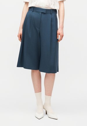 Kvinde iført knælange, mørkeblå plisserede culotter, hvide spidse hæle og cremefarvede strømper mod en ensfarvet baggrund.