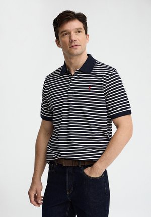 Mand iført navy og hvid horisontalt stribet polo med rød logo, mørke jeans og brun bælte, står med den ene hånd i lommen.