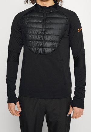 Mand iført en sort langærmet pullover med en quiltet forpanel og halvlukning med lynlås, med en lille orange Nike-logo på ærmet.