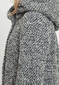 Donna che indossa un cappotto di lana grigio e bianco a motivo testurizzato con un ampio colletto, vista parziale che mostra la parte superiore del torso e ciocche di capelli biondi sciolti.