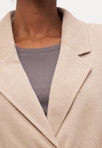 Blazer beige claro con tejido texturizado, solapas anchas y cuello con muesca, superpuesto sobre una camiseta de punto gris oscuro.