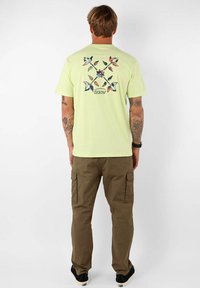 T-shirt en coton vert clair avec un design graphique multicolore au dos ; associé à un pantalon cargo marron et des chaussures noires.