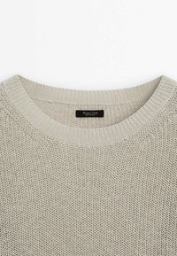 Pull en tricot gris clair avec un col rond, présentant un motif texturé et une petite étiquette de marque noire au niveau du col.