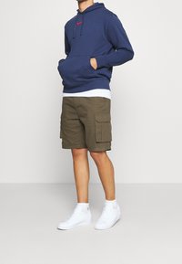 Navy hoodie with red Nike logo, olive cargo shorts with large side pockets, and white high-top sneakers with visible laces.