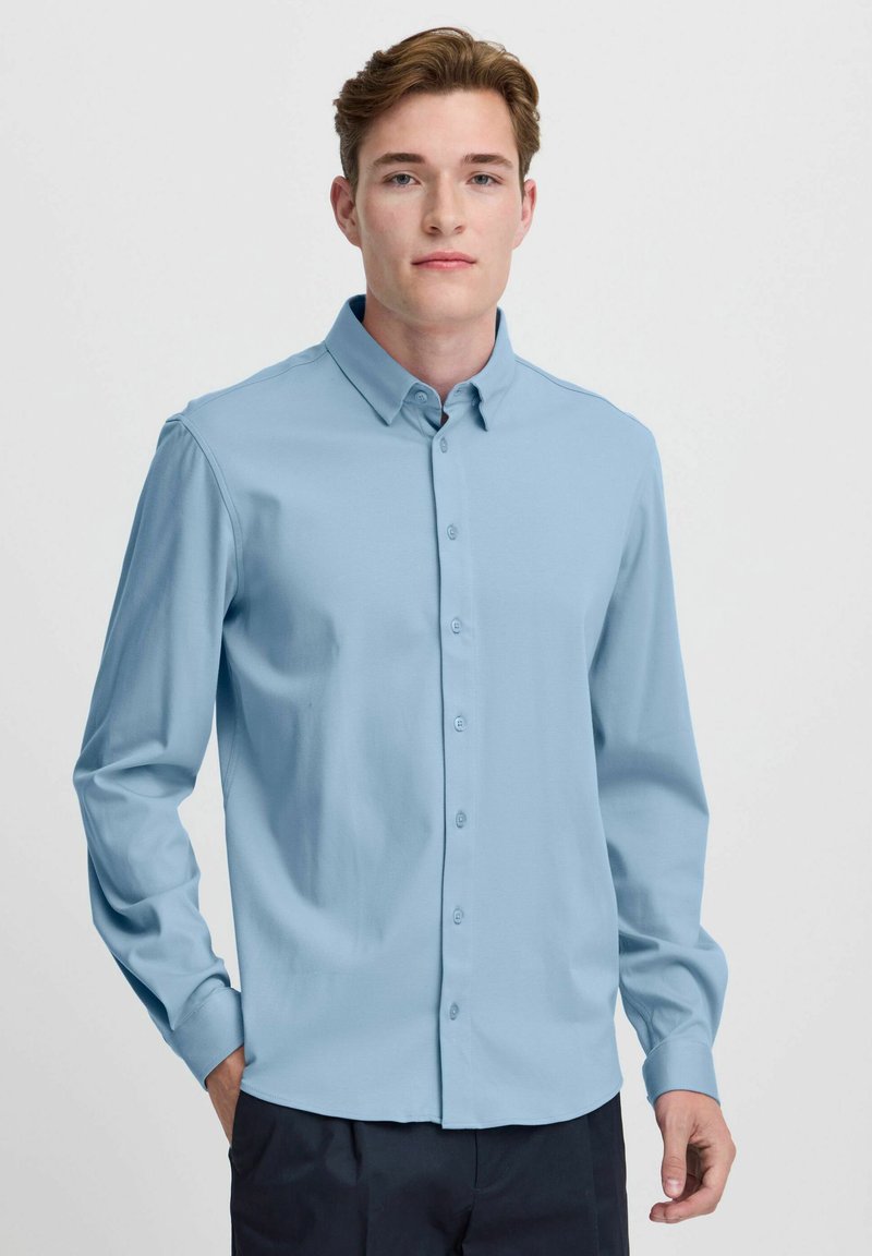 Camisa de manga larga azul claro con cuello abotonado. Presenta una textura suave y un cierre de botón estándar en la parte delantera.