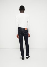 Jeans azul escuro com um corte ajustado, detalhes mínimos, e um patch de couro castanho na parte de trás da cintura; combinados com uma camisa de manga longa branca.