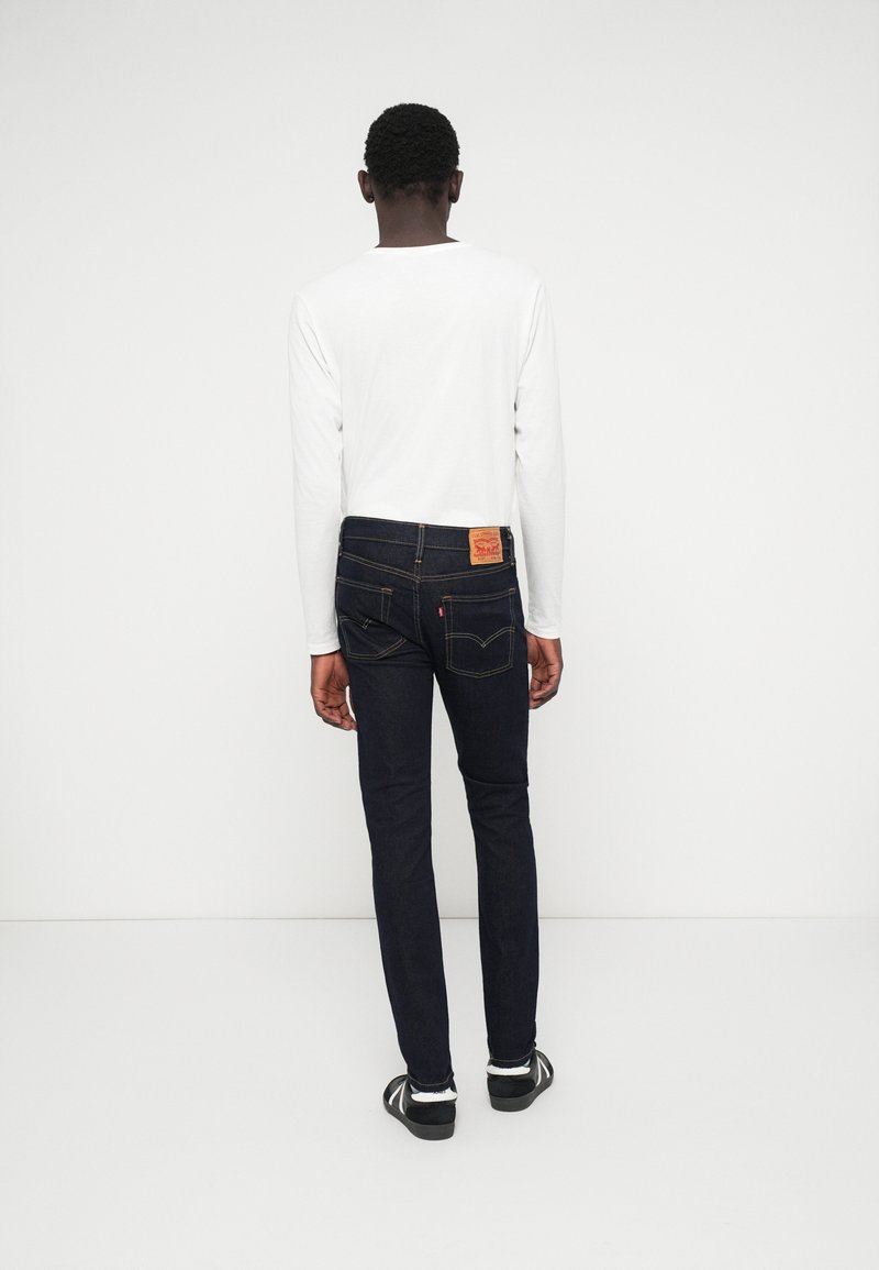 Jean bleu foncé avec une coupe ajustée, des détails minimaux et une petite pièce en cuir marron sur la ceinture arrière ; associé à une chemise blanche à manches longues.