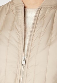 Närbild av en beige vadderad jacka som är delvis öppen över en vit stickad tröja.