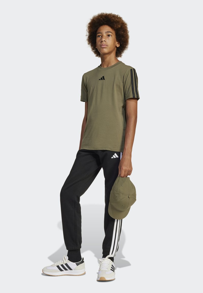 T-shirt verde oliva con strisce nere alle maniche, pantaloni jogger neri con logo Adidas bianco, sneaker bianche e nere, e un cappellino verde.