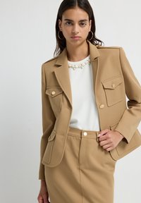 Mujer vestida con chaqueta beige a medida y falda a juego, blusa blanca con detalles de perlas y grandes pendientes de aro dorados, de pie frente a un fondo liso.