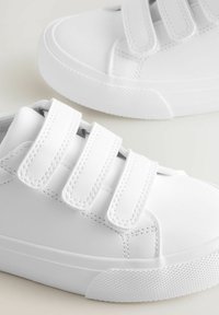 Weiße Sneaker mit drei Klettverschlüssen, ausgestattet mit einer glatten Oberfläche und einer strukturierten Sohle, vor einem hellen Hintergrund platziert.