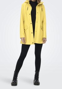 Gul vandtæt regnjakke med hætte, trykknapper og frontlommer. Båret over en sort top og sorte leggings, parret med sorte støvler.