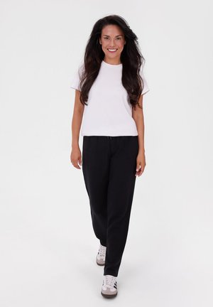 Giovane donna con lunghi capelli scuri sorride, camminando in avanti indossando una maglietta bianca, pantaloni neri e sneakers bianche su uno sfondo bianco uniforme.