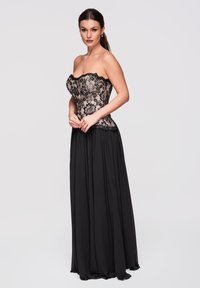 Femme aux cheveux foncés portant un corset en dentelle noire sans bretelles et une longue jupe noire fluide, posée devant un fond clair uni.