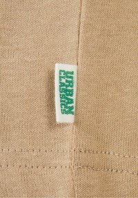 Tissu tricoté beige avec une petite étiquette blanche portant la mention "URBAN CLASSICS" en lettres vertes, cousue dans une couture.