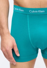 Άνδρας που φοράει τιρκουάζ εσώρουχα Calvin Klein boxer briefs, δείχνοντας τη λαστιχένια μέση με το λογότυπο και μέρος του αριστερού χεριού να ακουμπάει στο μηρό.