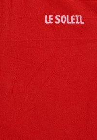 Fond en tissu rouge avec un texte blanc brodé "LE SOLEIL" en lettres majuscules près du coin supérieur droit.