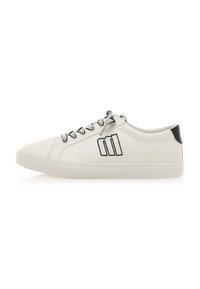 Zapatilla de cuero blanca con diseño de caña baja, acentos negros en el talón y un sutil detalle gráfico en el lado exterior. Cordones texturizados.