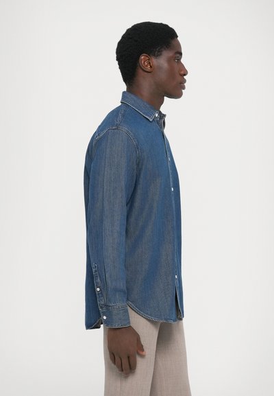 Chemise en denim bleu moyen, à manches longues, boutonnée à l'avant, col standard et ourlet arrondi. Tissu texturé avec de subtils détails de décoloration.