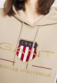 Felpa beige con logo e scudo ricamati, con strisce rosse e bianche. Coulisse con dettagli in metallo e una piacevole texture.