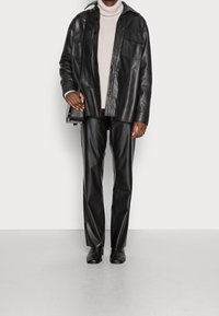 Svart skjortjacka i läder med två bröstfickor, bärs över en ljusgrå polotröja, kombinerad med matchande svarta läderbyxor och ankelboots.