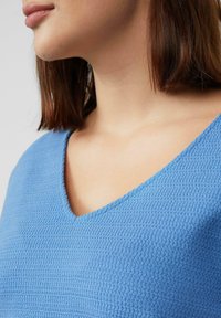 Blauwe gestructureerde top met een V-halsontwerp, gemaakt van een geweven stof. Close-upweergave benadrukt de details van de stiksels en het stofpatroon.