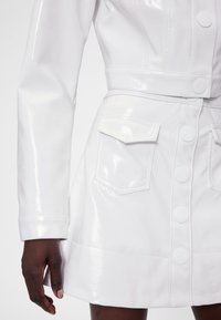 Bela, sijo umetna usnjena obleka z oprijetim pasom, dolgimi rokavi, dvema sprednjima žepoma in zapiranjem na gumbe spredaj.