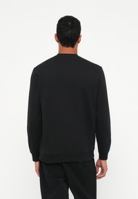 Hombre de pie de espaldas, vistiendo un sudadera negra de manga larga y pantalones negros, contra un fondo blanco.