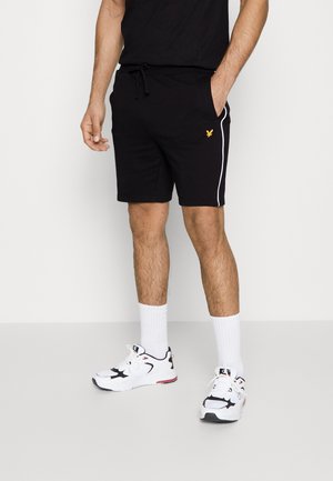 Man in zwarte sportbroek met witte zijstreep en klein geel logo, witte sportsokken en witte sneakers, staand tegen een effen achtergrond.