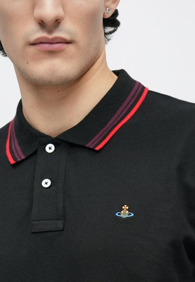 Posnetek iz bližine moškega, ki nosi črno polo majico z rdeče in vijolično črtastim ovratnikom ter z manjšim vezenim pisanim logotipom na prsih.