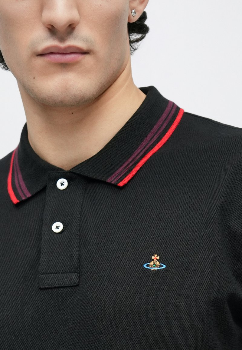 Nærbillede af en mand iført en sort polo med en rød og lilla stribet krave og en broderet lille farverig logo på brystet.