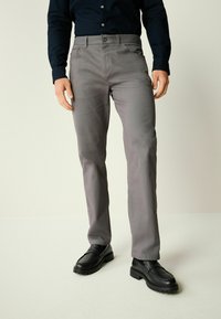 Mand iført slim-fit grå bukser, sorte læder loafers og en sort langærmet skjorte, står på en lys baggrund.