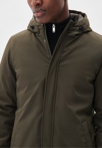 Man i en olivgrön huvjacka som är zipad över en svart höghalspullover med en silverdragkedja synlig.