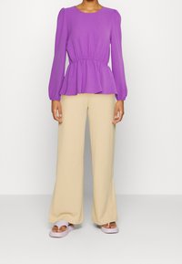 Top a peplo viola con maniche arricciate, abbinato a pantaloni a gamba larga beige chiaro e scarpe slip-on viola chiaro. Texture morbide, design casual.