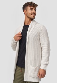 Cardigan lavorato a maglia color beige chiaro con colletto a scialle e apertura frontale. Presenta polsini a costine e una tasca applicata sul davanti. Texture morbida.