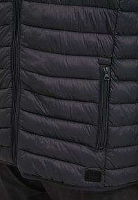 Svart puffervest med quiltet konstruksjon, med en glidelåslomme på siden og en liten logotag i kanten. Glatt tekstur.