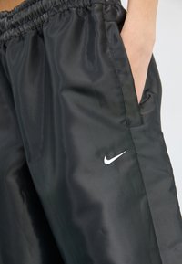 Melni sporta bikses, izgatavotas no gludas, spīdīgas vielas, ar elastīgu jostasvietu un sānu kabatām. Piebilde: neliels balts Nike logo.