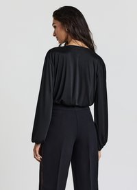 Schwarze Satinbluse mit langen Ärmeln und lockerem Schnitt, kombiniert mit hochtaillierten schwarzen Hosen mit seitlichen Spitzenbesätzen.