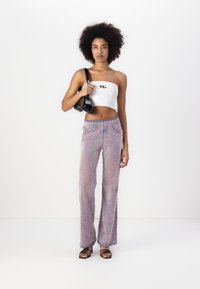 Bílý žebrovaný crop top s logem, doplněný volnými džíny s flared nohavicemi v růžových odstínech. Modelka drží černou kabelku. Vzhled doplňují sandály na tenkých popruzích.