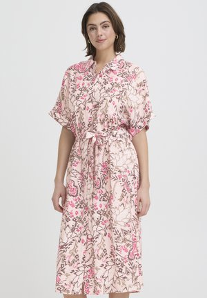 Kobieta ubrana w różową sukienkę midi w kwiaty z krótkimi rękawami, zapinana na guziki z kołnierzykiem i przewiązana w talii, stojąca na jasnym, jednolitym tle.