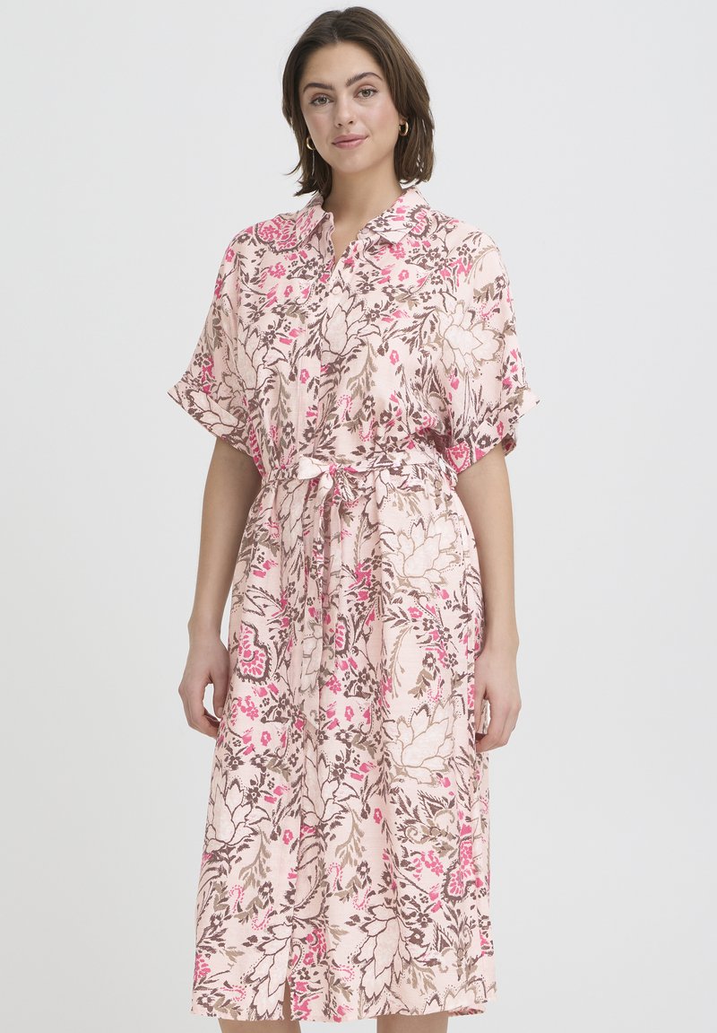 Mujer con un vestido midi floral rosa con mangas cortas, botones en el frente, cuello y cintura atada, de pie contra un fondo claro y liso.