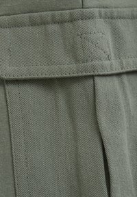 Zaļš, teksturēts audums ar izteiktu audumu, kas rotāts ar sīku diegu rakstu un salocītu jostu ar smalku logo.